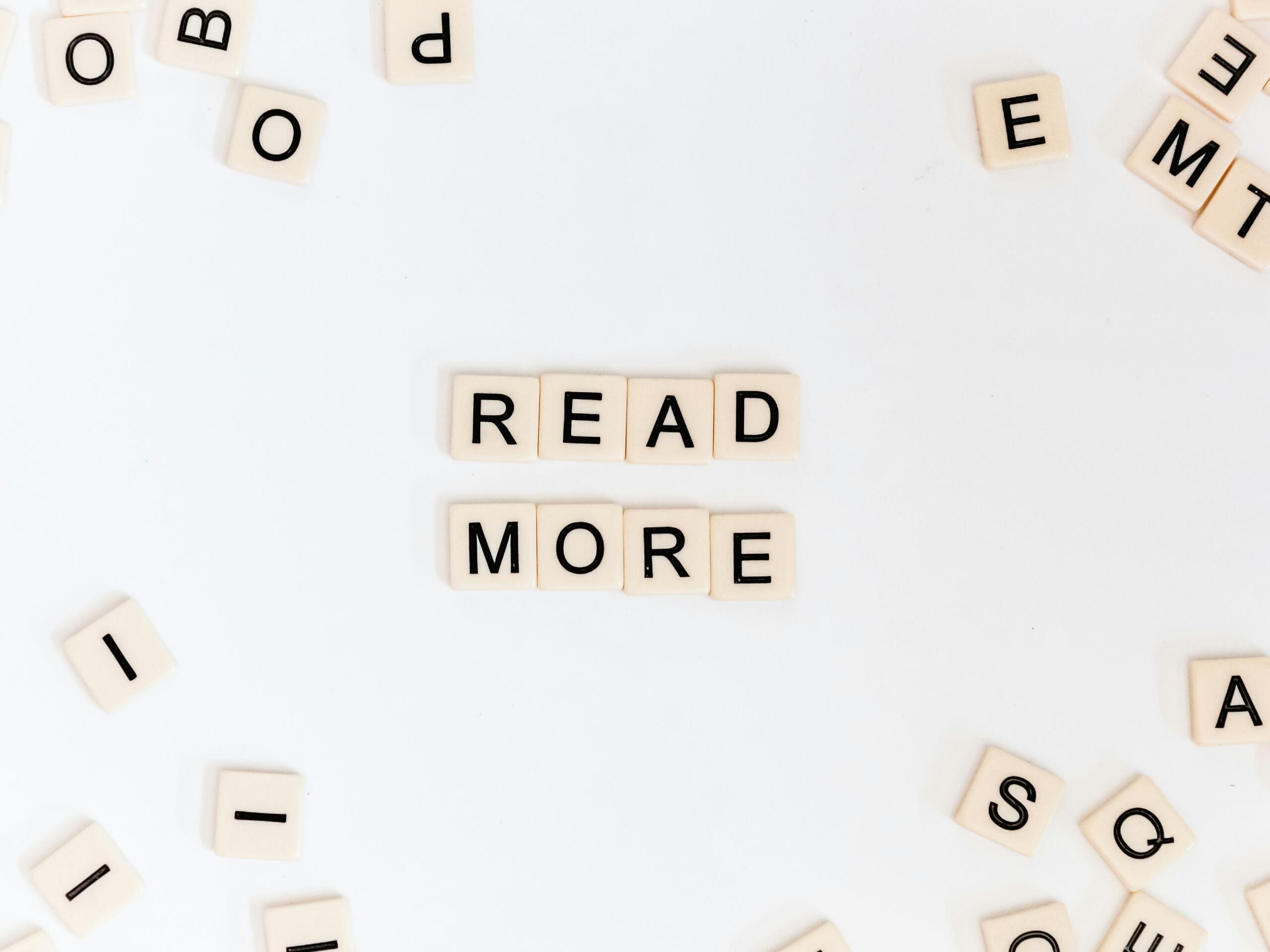 Fichas de Scrabble dispuestas para formar la frase &laquo;READ MORE&raquo; (LEER M&Aacute;S) con letras dispersas sobre una superficie blanca.
